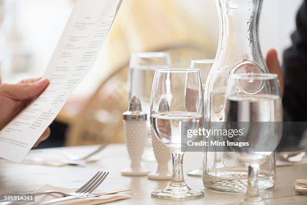 close up of wine and water glasses and place settings at a table in a restaurant. a persons hand holding the menu. - carafe stock pictures, royalty-free photos & images