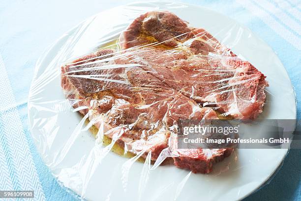 two pork collar steaks in olive oil marinade on a white plate under cling film - fresh meat film stock pictures, royalty-free photos & images