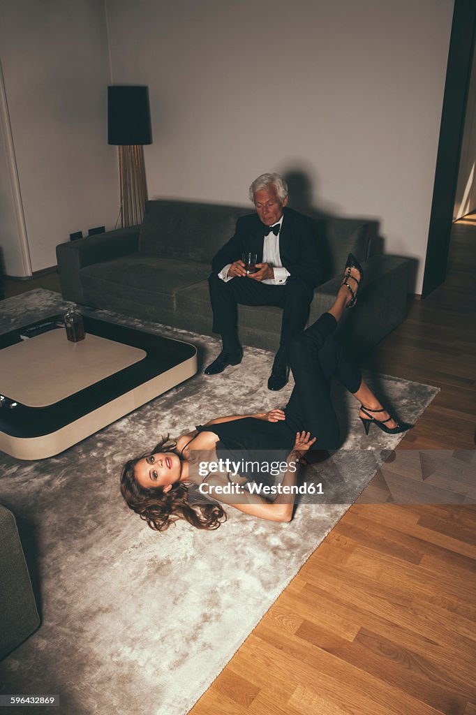 Senior man with drink on sofa looking at young woman lying on carpet