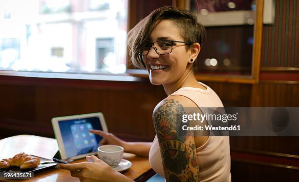 portrait of tattooed young woman sitting in a coffee shop using digital tablet - tattoo shoulder stock-fotos und bilder