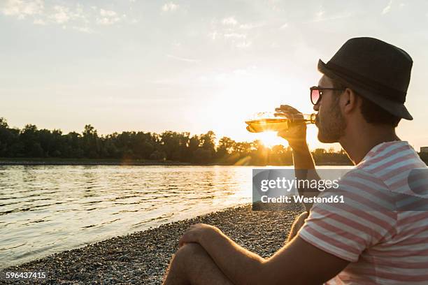 young man drinking beer at the riverside at sunset - riverbank stock-fotos und bilder