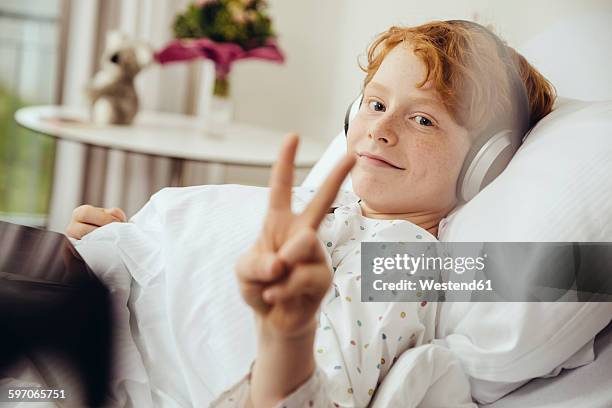 sick boy lying in hospital making victory sign, wearing head phones - children's hospital imagens e fotografias de stock