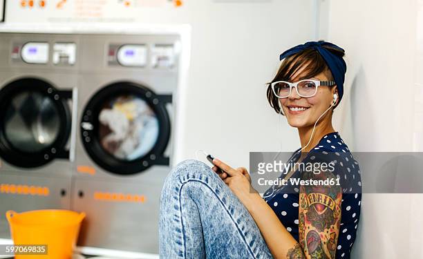 portrait of tattooed young woman hearing music with earphones in a launderette - tattoo shoulder stock-fotos und bilder