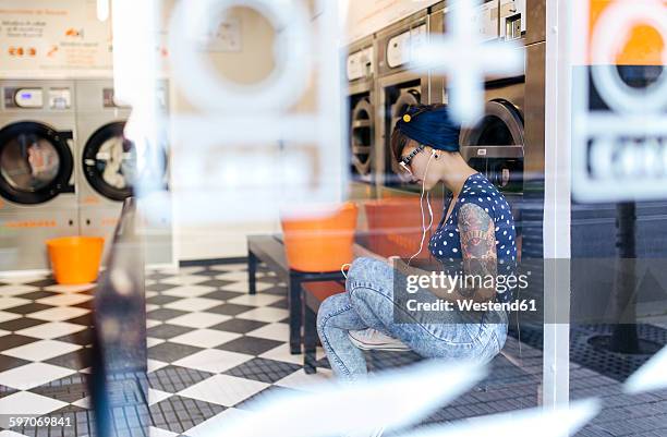 tattooed young woman hearing music with earphones in a launderette - dry cleaners stock pictures, royalty-free photos & images