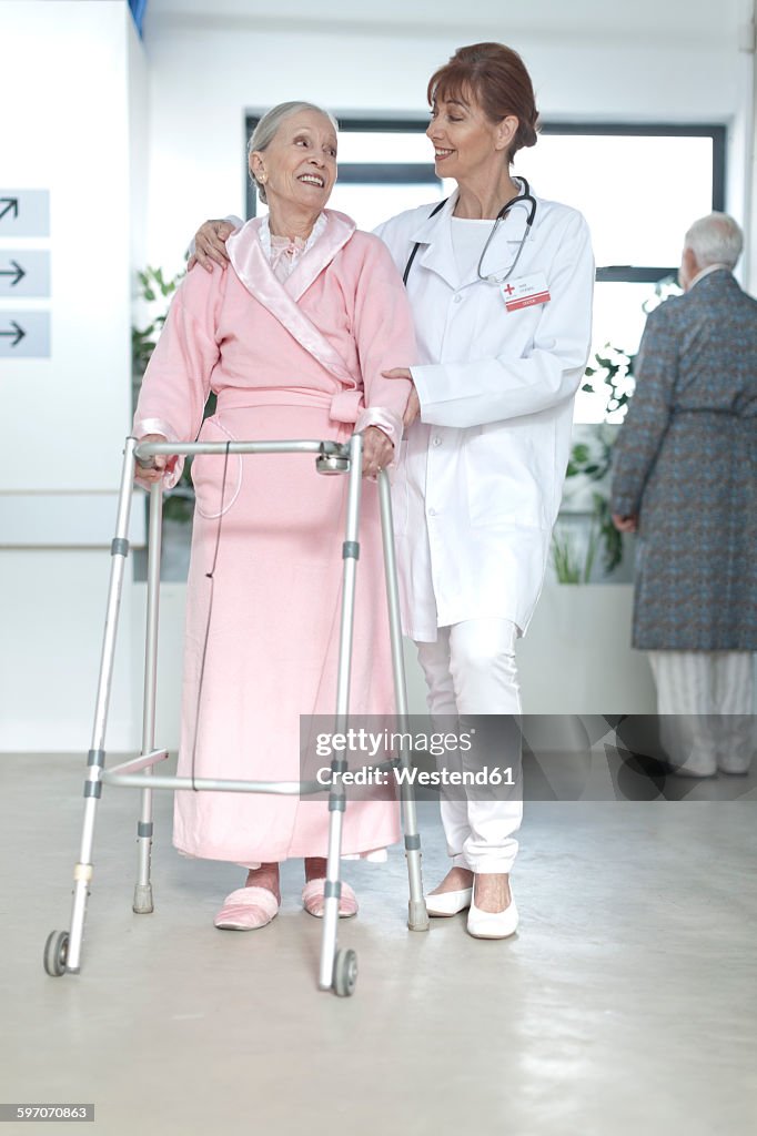 Doctor leading elderly patient with walking frame on hospital floor