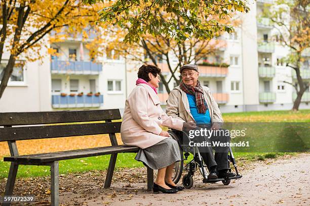 senior woman sitting on bench next to husband in wheelchair - bench imagens e fotografias de stock