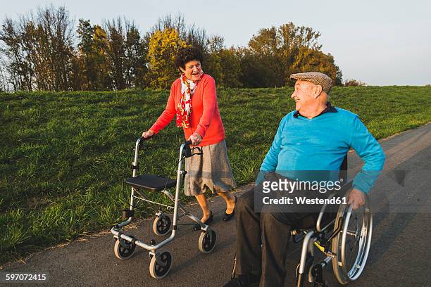 smiling senior couple with wheelchair and wheeled walker on a path - rollator stock-fotos und bilder