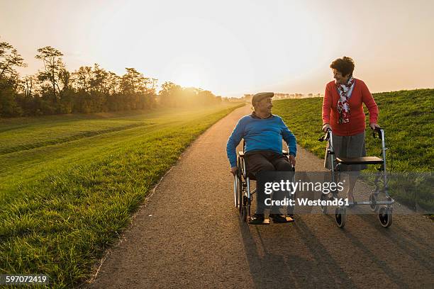smiling senior couple with wheelchair and wheeled walker on a path - rollator stock-fotos und bilder