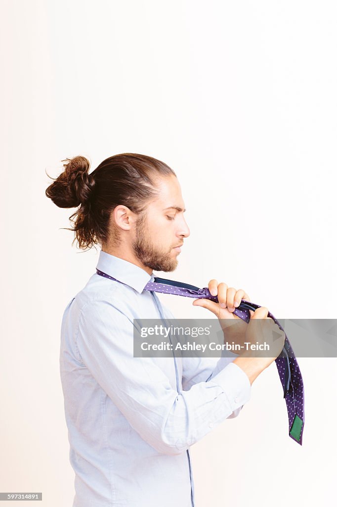 Young man with hair in bun, getting dressed, tying neck tie