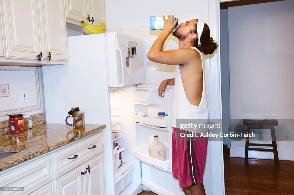 Young man standing next to open fridge, drinking milk from carton
