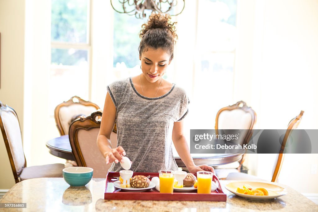 Young woman at home preparing breakfast tray, looking down holding egg