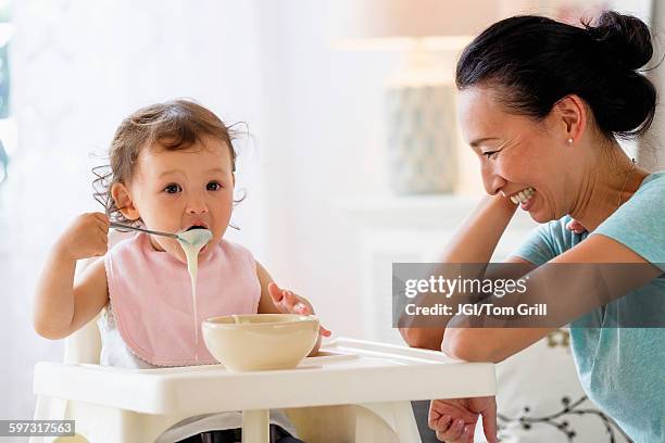 mother watching baby daughter eat in high chair - baby eating yogurt stockfoto's en -beelden