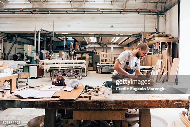 young man in carpentry workshop looking down sanding skateboard - carpintería fotografías e imágenes de stock