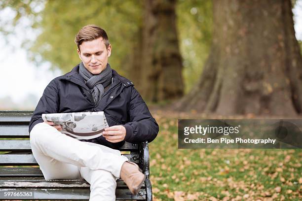 young man reading newspaper on park bench - junger mann liest zeitung stock-fotos und bilder