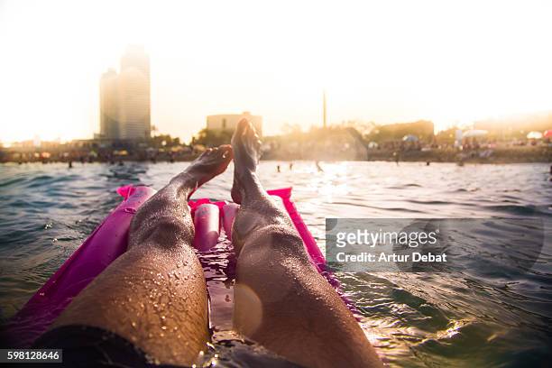 guy swimming on pink inflatable bed pad on mediterranean sea from personal perspective with the skyline of the barcelona city on sunset during summer time without stress and relaxing times. - la barceloneta ストックフォトと画像
