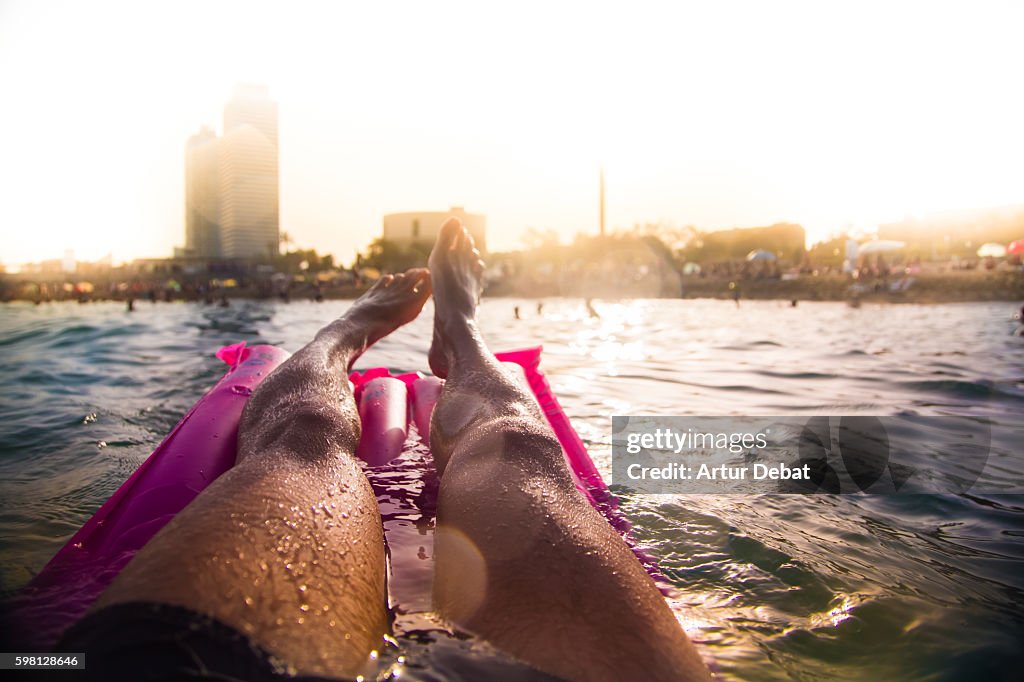 Guy swimming on pink inflatable bed pad on Mediterranean Sea from personal perspective with the skyline of the Barcelona city on sunset during summer time without stress and relaxing times.