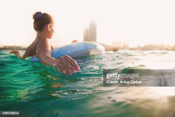 Girl Swimming On Inflatable Ring Pad On Mediterranean Sea With The Skyline Of The Barcelona City On Sunset During Summer Time Without Stress And Relaxing Times, Foto de stock