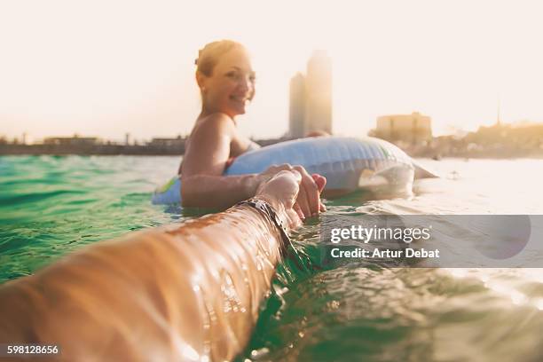 girl swimming on inflatable ring pad on mediterranean sea with boyfriend taking his hand and taking picture from personal perspective with the skyline of the barcelona city on sunset during summer time without stress and relaxing times. follow me. - hot spanish women ストックフォトと画像