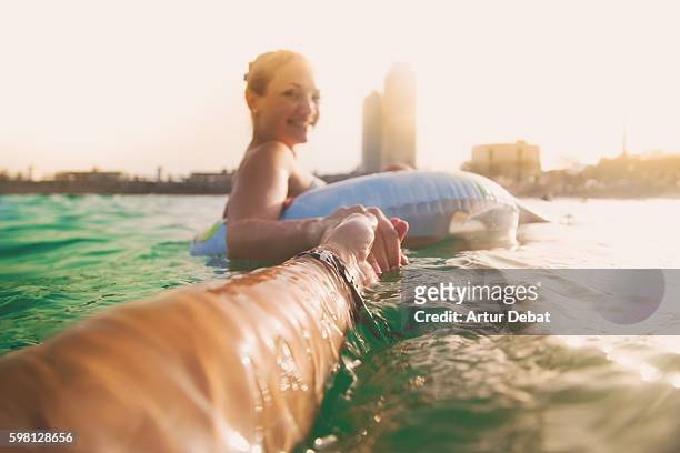 girl swimming on inflatable ring pad on mediterranean sea with boyfriend taking his hand and taking picture from personal perspective with the skyline of the barcelona city on sunset during summer time without stress and relaxing times. follow me. - europe asian culture stockfoto's en -beelden