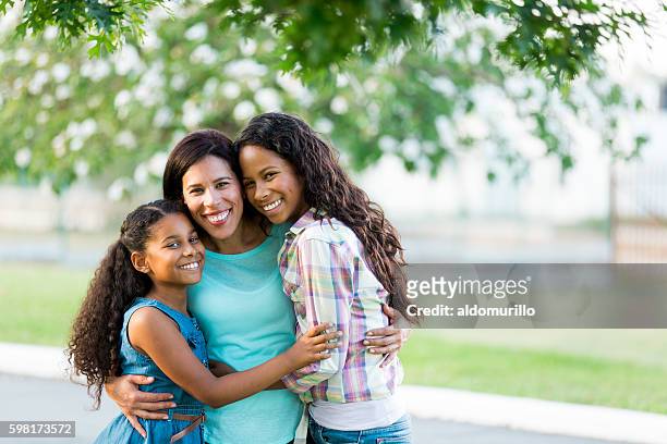 madre e figlie che si abbracciano strettamente e sorridono alla telecamera - family with two children foto e immagini stock