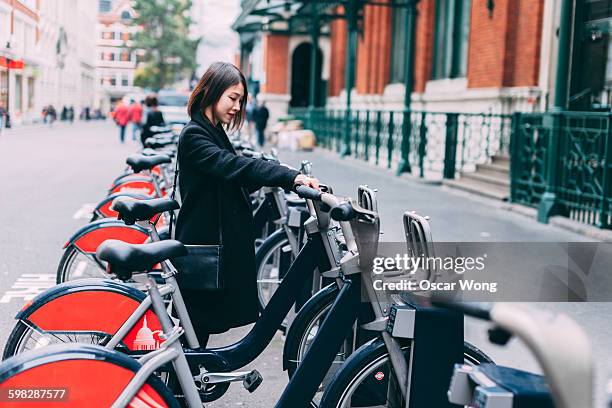 young woman renting bicycle on street in london - bicycle rental stock pictures, royalty-free photos & images