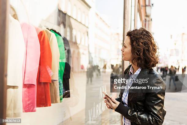 businesswoman doing window shopping while holding mobile phone in city - window shoppen stockfoto's en -beelden