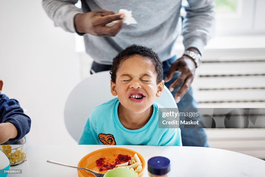 Boy making face while having food at table with father standing in background