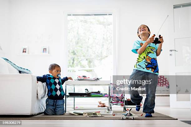 boy looking at happy brother holding remote of model airplane at home - radiografische afstandsbediening stockfoto's en -beelden