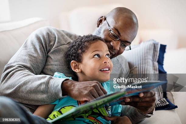 smiling boy looking at father while reading picture book at home - famille avec des lunettes de vue photos et images de collection
