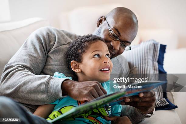 smiling boy looking at father while reading picture book at home - familly glasses stockfoto's en -beelden