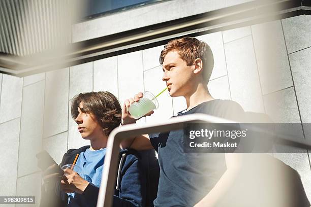 boy looking away while drinking juice with friend holding smart phone outdoors - drinking straw photos et images de collection