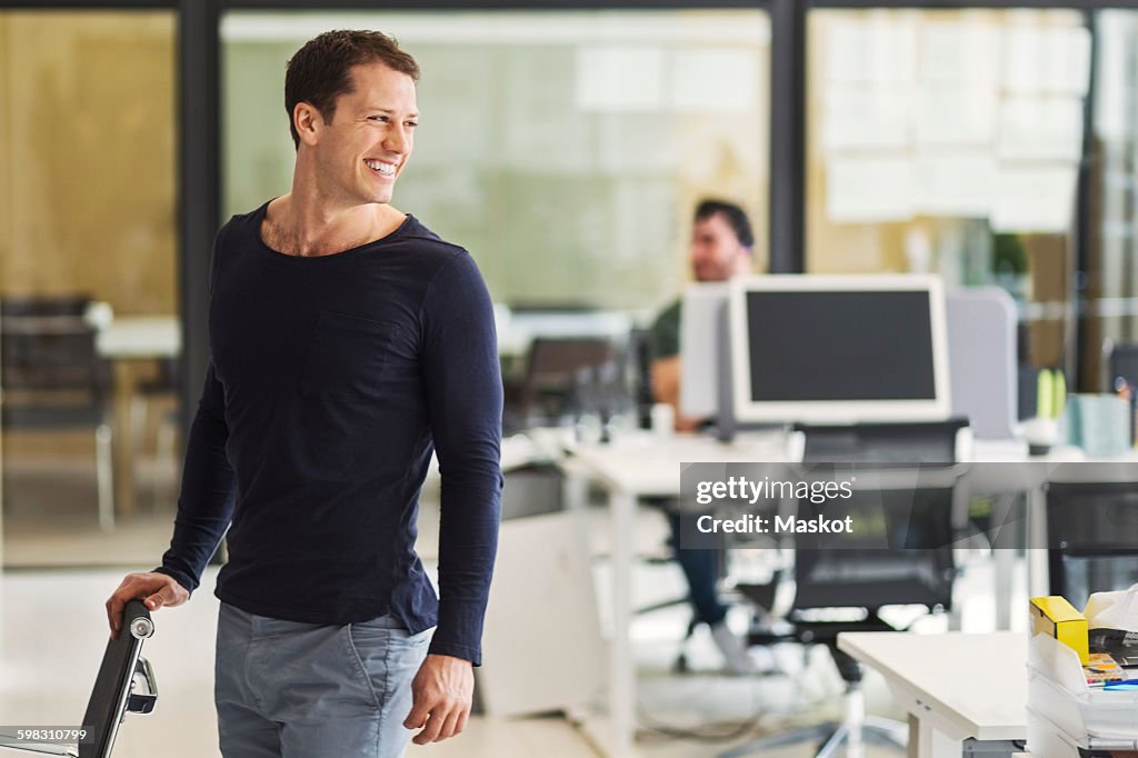 Happy mid adult businessman looking away with colleague in background at office