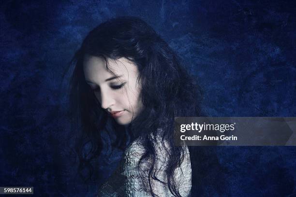 pale young woman with tousled long dark hair looks down sadly, textured dark blue background, studio shot - pale complexion fotografías e imágenes de stock