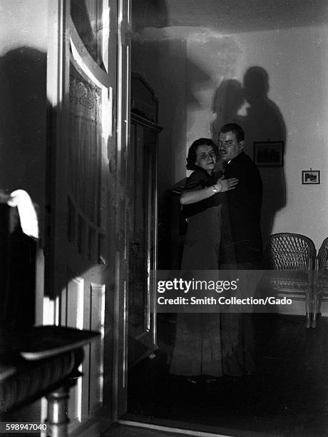 In a dark, starkly lit room, a couple dances in the middle distance, with their shadows cast on the wall behind them, Hungary, 1929. Digitized from a...