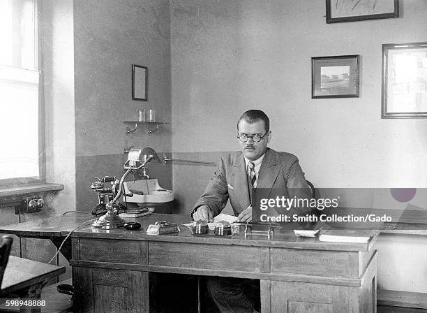 Man with short hair, round spectacles, a conservative suit and a stern facial expression sits behind a large wooden desk in his office, with a small...