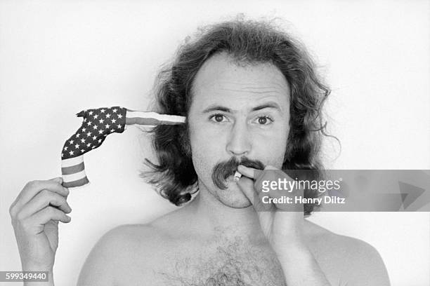 David Crosby holds a toy gun to his head while smoking a marijuana cigarette. The gun is made of cloth soft sculpture and decorated with stars and...