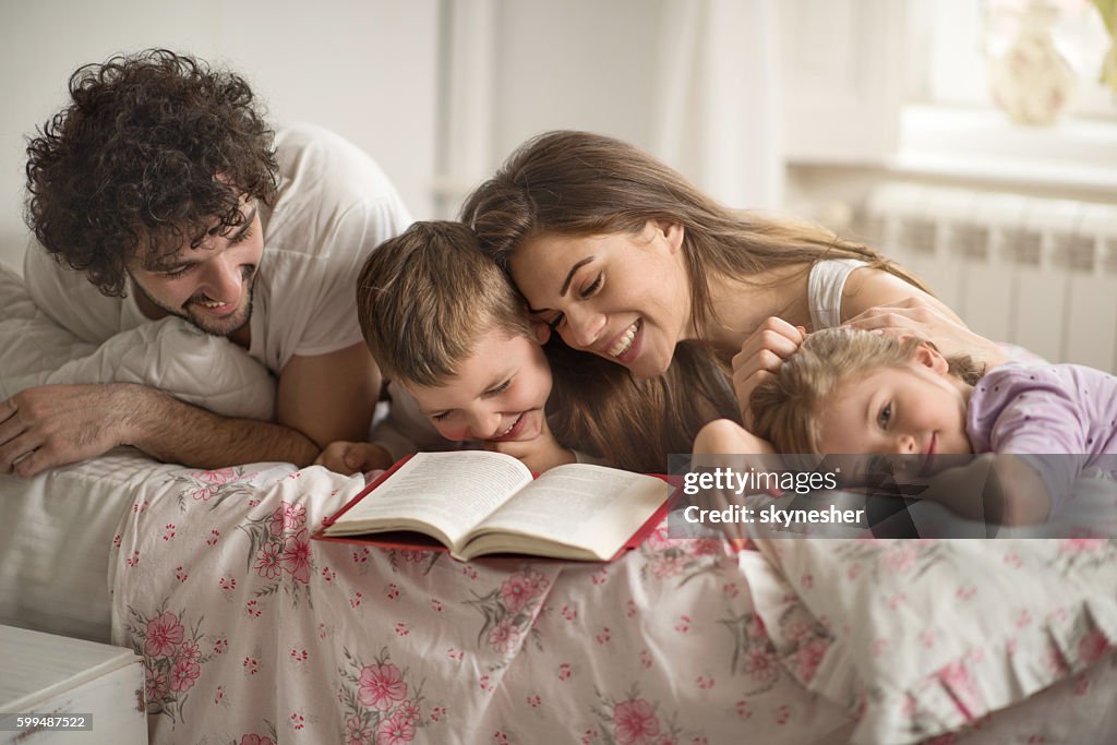 Joyful family reading a book and laughing in the bedroom.