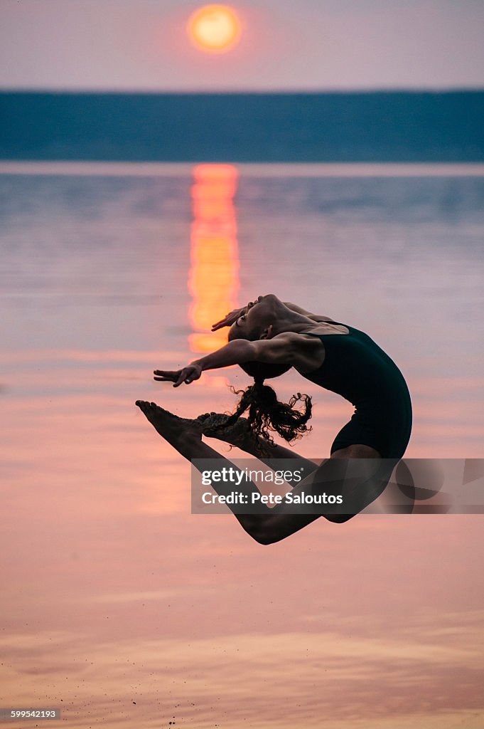 Side view of girl by ocean at sunset, leaping in mid air bending backwards