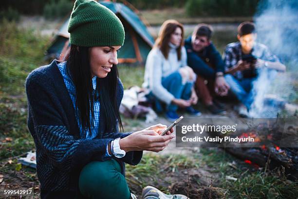 young woman using mobile phone while friends sitting by bonfire at campsite - remote location cell phone stock pictures, royalty-free photos & images