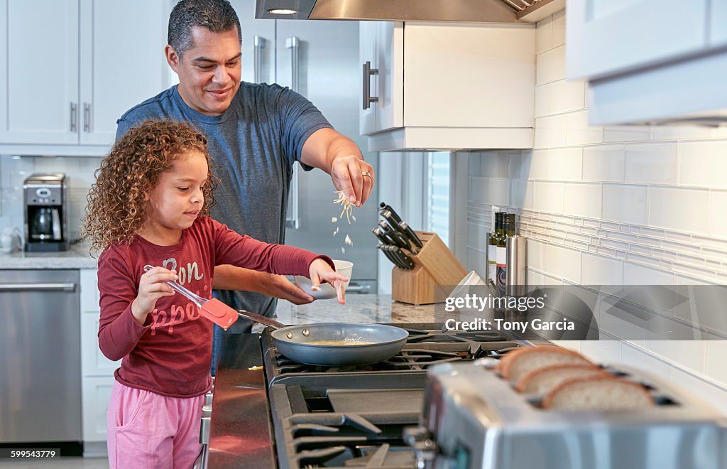 Father helping daughter cook on hob in kitchen, sprinkling cheese