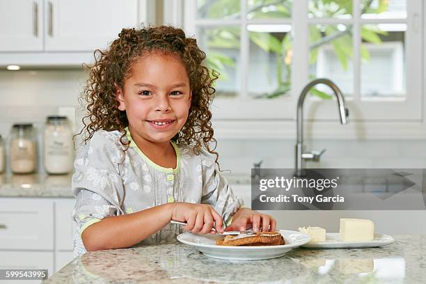 girl at kitchen counter spreading butter on toast looking at camera smiling - untar de mantequilla fotografías e imágenes de stock