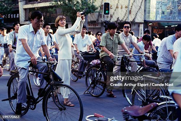 Linda Evangelista standing amid bicyclists on a street in Shanghai, wearing an ivory ribbed turtleneck by Joseph Tricot and a skirt by Calvin Klein...