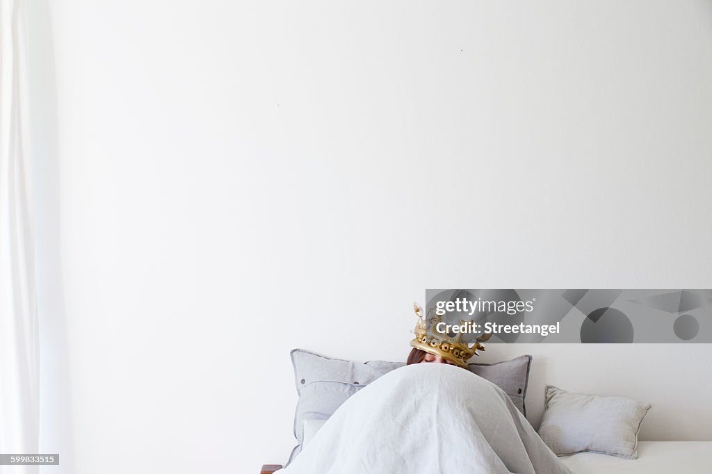 Mature woman in bed underneath quilt wearing golden crown