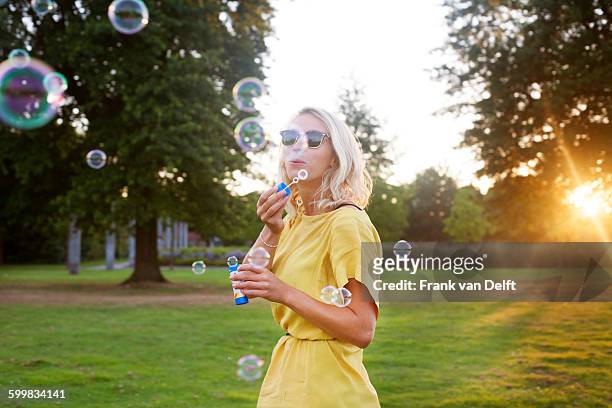 portrait of young woman wearing yellow dress blowing bubbles in park at sunset - bubble wand foto e immagini stock