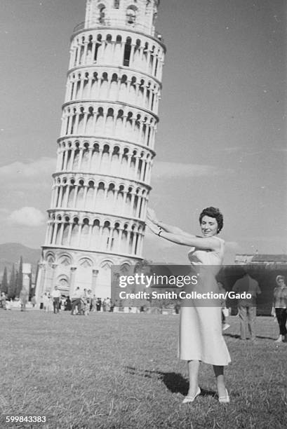 Leaning Tower of Pisa, a young female tourist in a dress standing in a field, smiling and pretending to hold up the tower, other tourist groups...