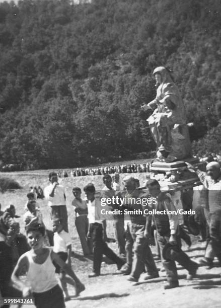 Feast of San Gennaro, a large group of villagers carrying a statue of Saint Gennaro along a dirt path, several men holding wooden staves and...