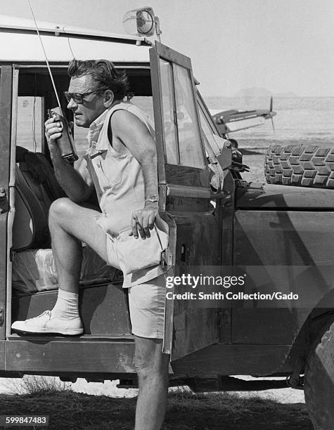 William Holden, movie still, standing with one leg up on the door of an off-road vehicle, using a walkie talkie, an airplane in the background, 1969.