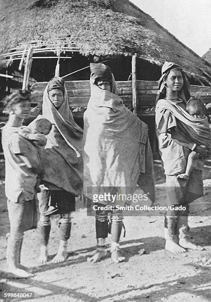 Padaung tribe members, standing near straw huts and modeling cold weather clothing, Myanmar , 1922.