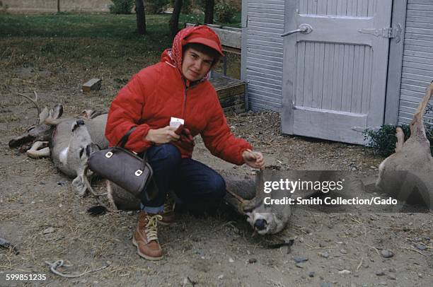 Woman wearing high visibility red vest, kneeling, holding the antlers of a deer carcass in one hand, with a purse draped over her opposite arm, on a...