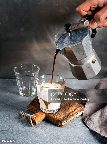 glass of coffee with ice cream on rustic wooden board. drink is poured from steel italian moka pot held by man's hand - moka pot stockfoto's en -beelden